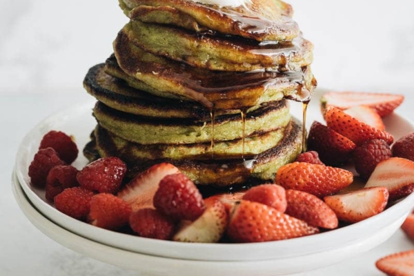 Close up of a stack of matcha pancakes with strawberry topping, whipped cream, and syrup