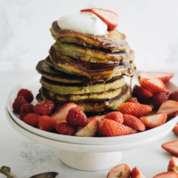 Squared picture of one stack of matcha pancakes with strawberry topping, whipped cream, and syrup