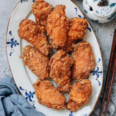 A plate of golden, crispy Chinese fried chicken wings served on a white platter with blue patterns, with a bowl of salt and chopsticks on the side.