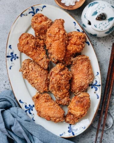 A plate of golden, crispy Chinese fried chicken wings served on a white platter with blue patterns, with a bowl of salt and chopsticks on the side.