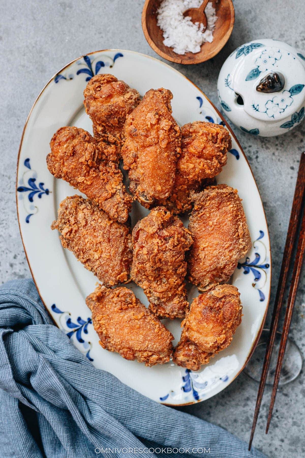 A plate of golden, crispy Chinese fried chicken wings served on a white platter with blue patterns, with a bowl of salt and chopsticks on the side.