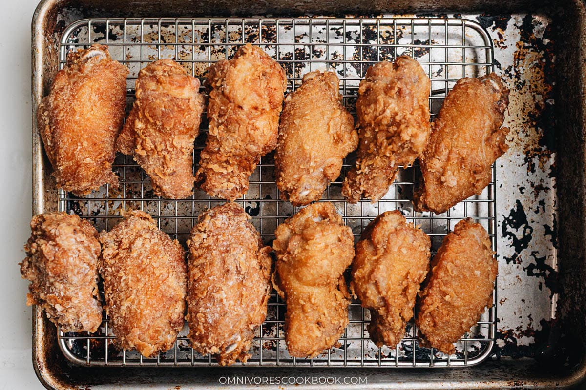 Freshly fried chicken wings resting on a wire rack over a baking sheet to drain excess oil after first frying.
