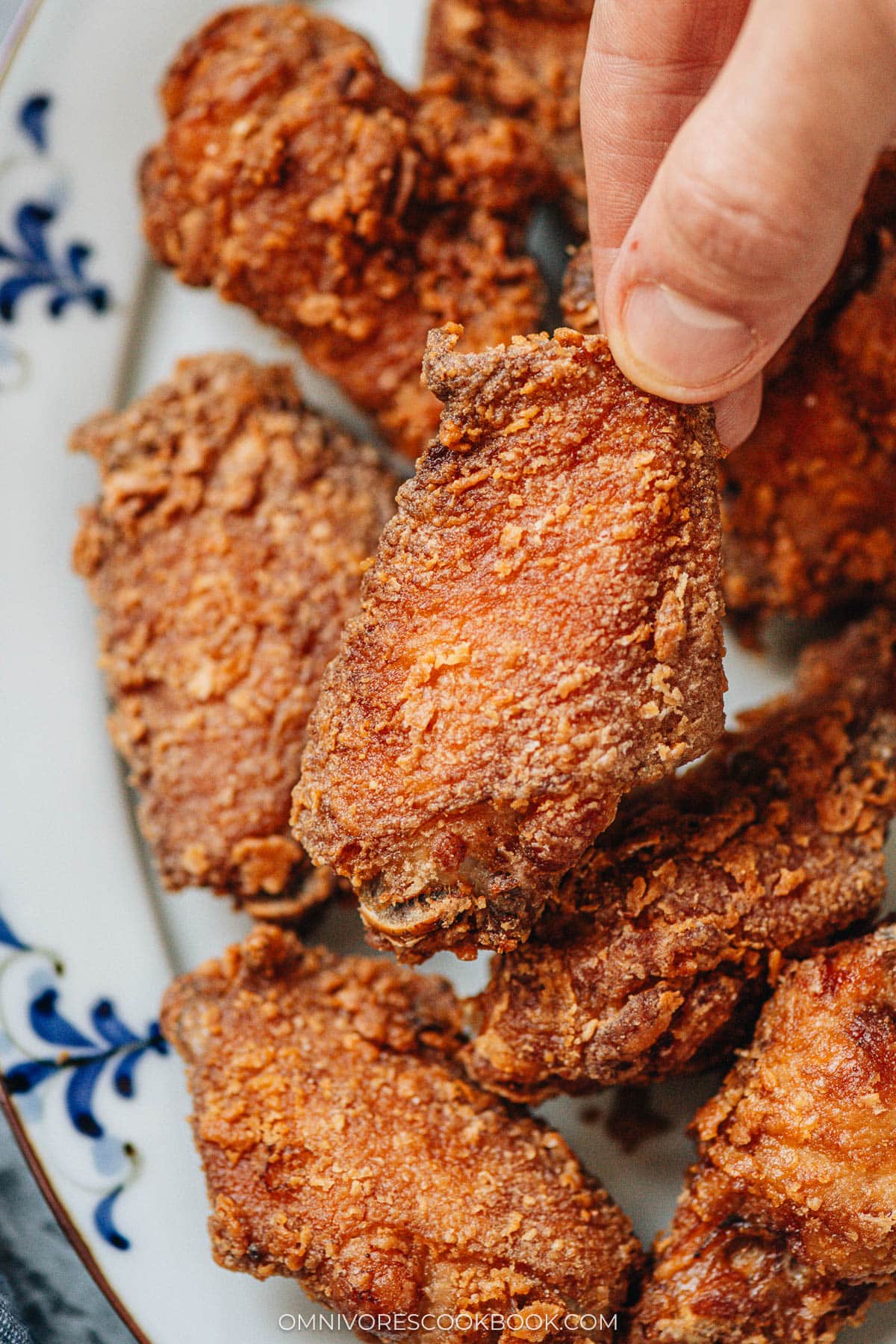 A hand picking up a freshly fried chicken wing, showing the crisp and craggy surface up close.