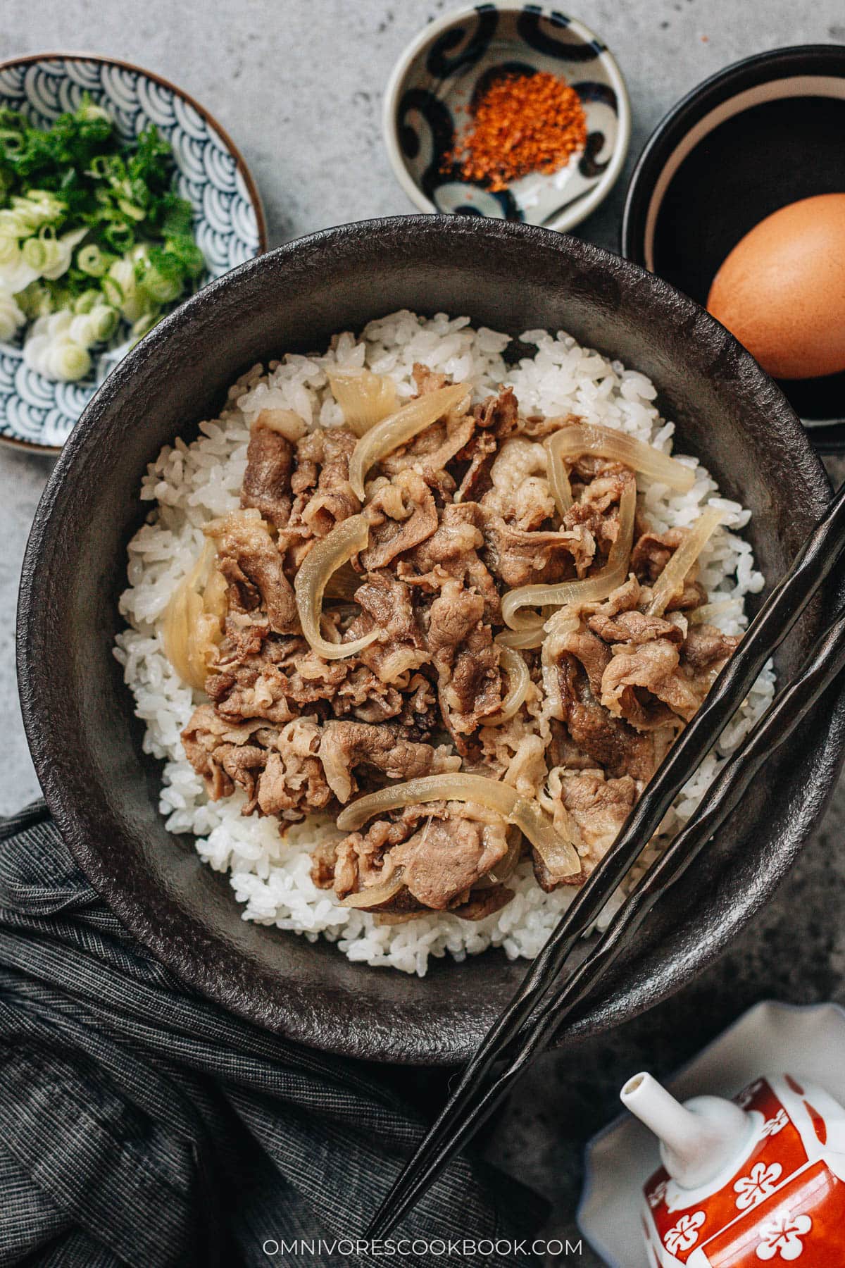 A bowl of gyudon with thinly sliced beef and onions simmered in savory sauce served over steamed rice, with small dishes of scallions, shichimi togarashi, and a raw egg beside it.