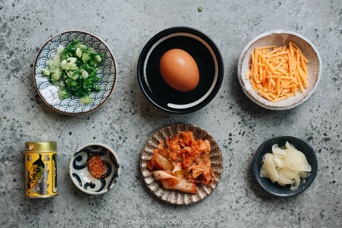 Small dishes of gyudon toppings and condiments arranged on a gray surface, including sliced scallions, a whole egg, shredded cheese, kimchi, pickled ginger, and shichimi togarashi.