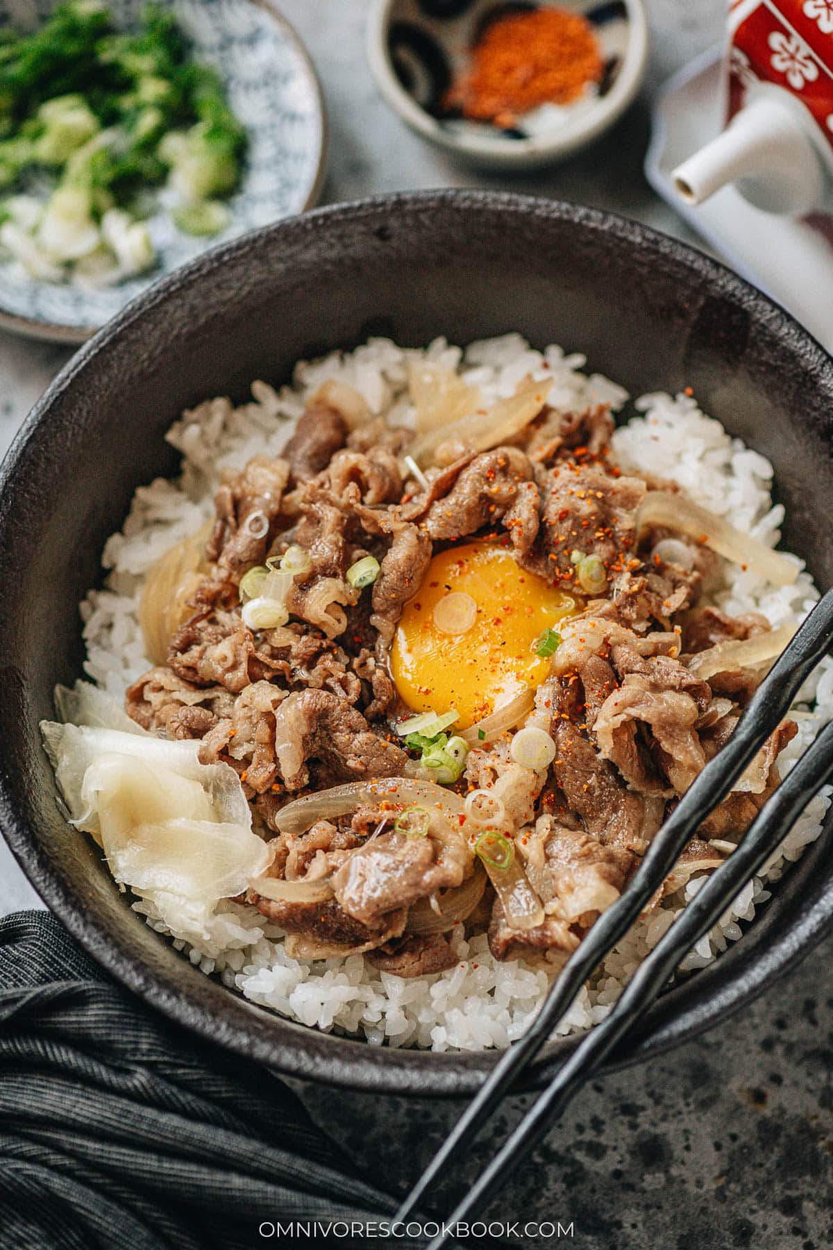 Gyudon topped with a raw egg yolk, sliced scallions, and a sprinkle of shichimi togarashi in a black bowl over rice, with a side of pickled ginger.