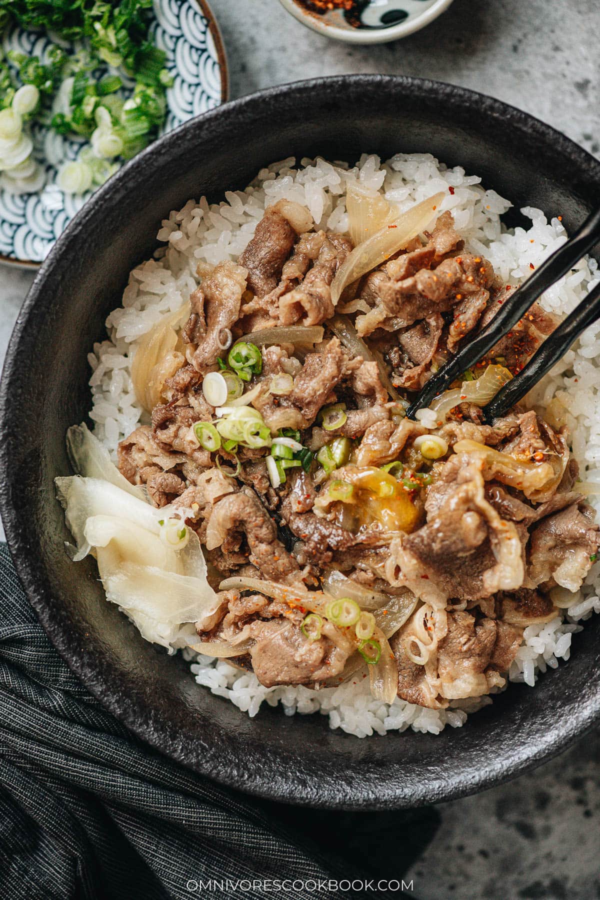 Chopsticks mixing gyudon topped with a soft raw egg yolk, beef, onions, and scallions over a bed of rice.