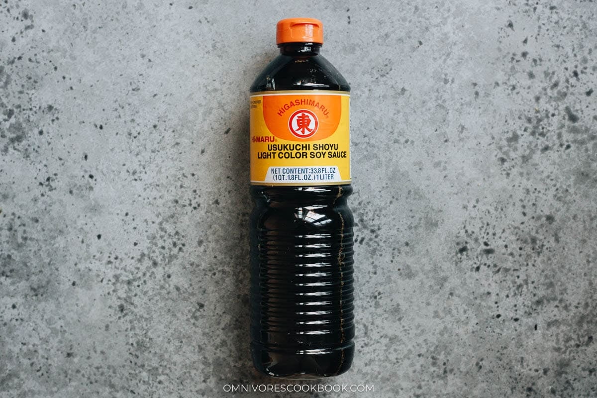 Bottle of Higashimaru usukuchi shoyu (light color soy sauce) placed on a gray countertop. Sliced onions simmering with kombu in broth inside a skillet.
