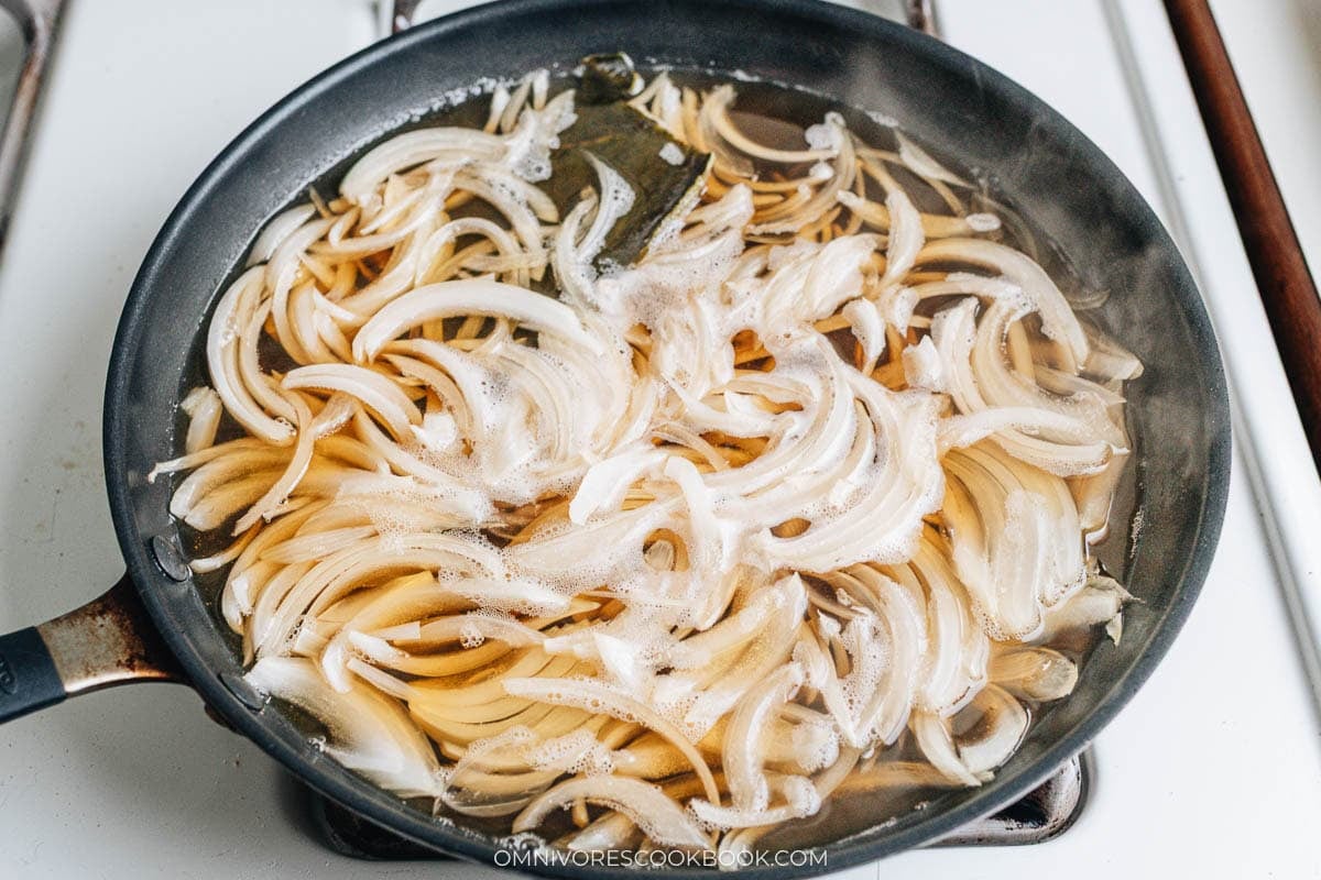 Sliced onions simmering with kombu in broth inside a skillet.
