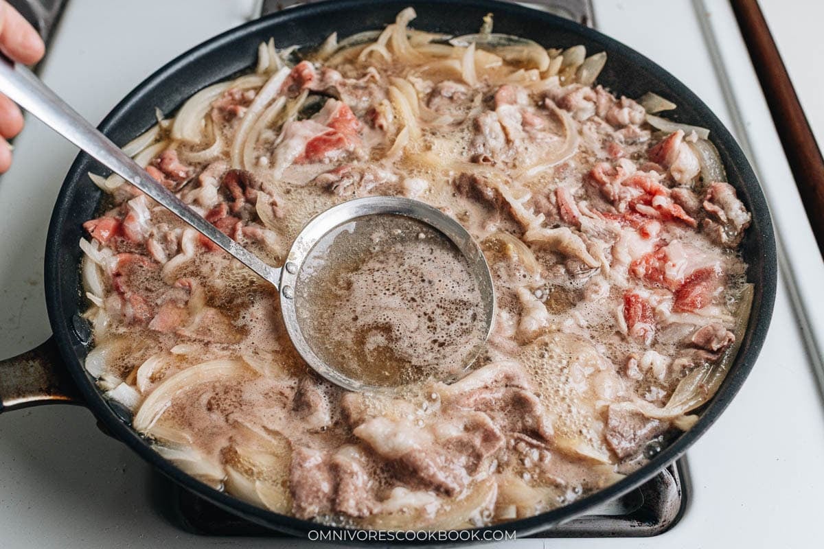 Thinly sliced beef simmering with onions in broth, with foam being skimmed off using a ladle.