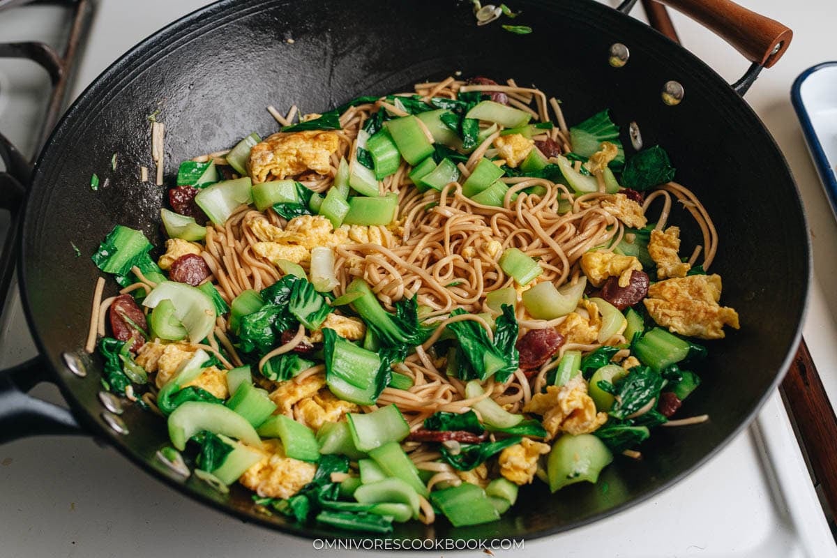 Wok filled with stir-fried noodles, bok choy, eggs, and sausage before the final toss.