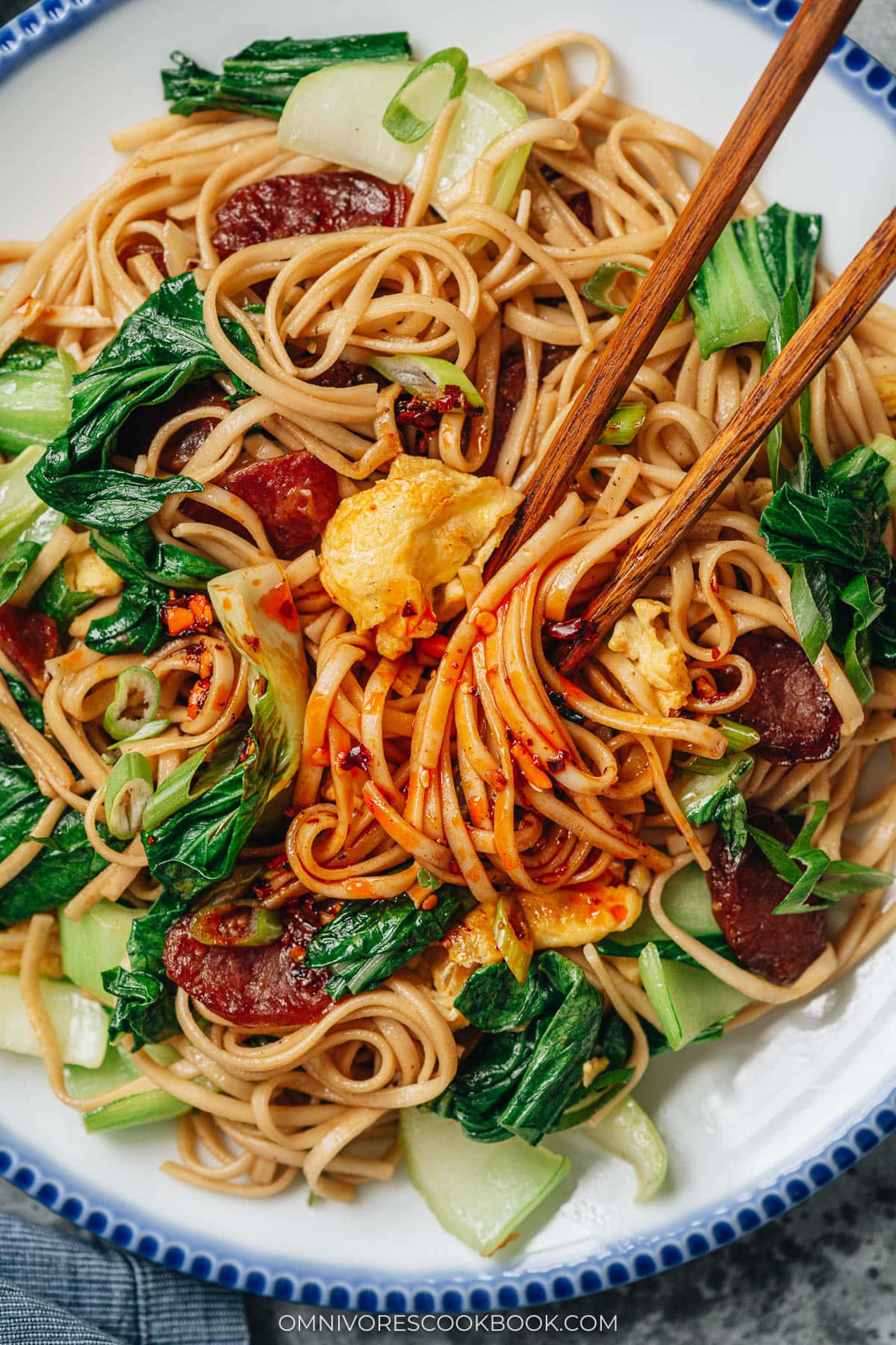 Chinese fried noodles drizzled with chili crisp, served with greens, sausage, and eggs for a spicy noodle dish.