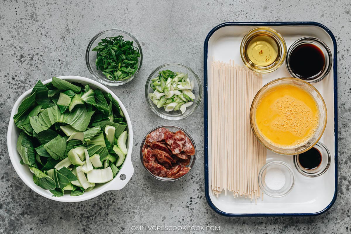 Ingredient layout including bok choy, Chinese sausage, eggs, scallions, oil, soy sauce, and dried noodles.