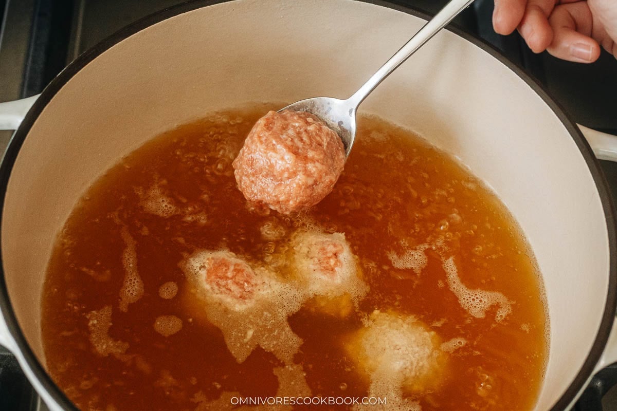 A hand shapes a pork meatball with a spoon and gently drops it into a pot of simmering broth