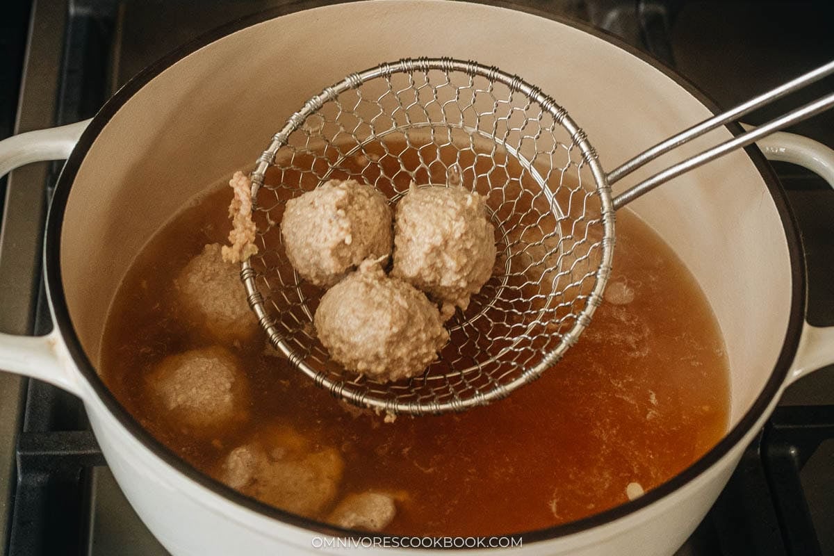 Cooked pork meatballs are lifted from a pot of simmering broth using a wire skimmer