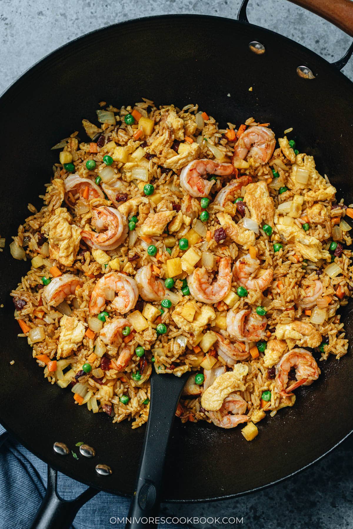A wok filled with Thai pineapple fried rice with shrimp, eggs, peas, and diced vegetables, stirred with a black spatula.