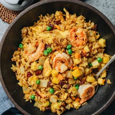 A bowl of pineapple fried rice topped with shrimp, peas, and chunks of pineapple, with a spoon resting inside.