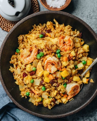 A bowl of pineapple fried rice topped with shrimp, peas, and chunks of pineapple, with a spoon resting inside.