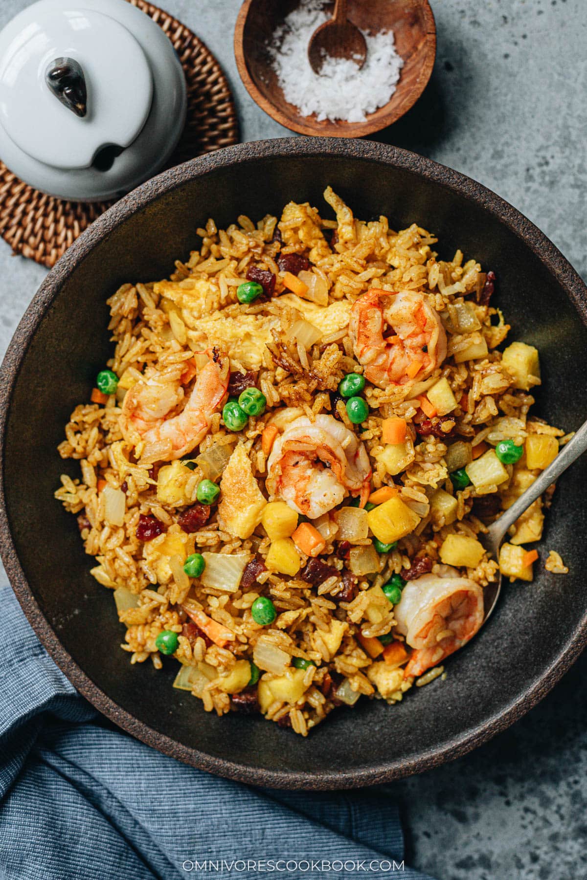 A bowl of pineapple fried rice topped with shrimp, peas, and chunks of pineapple, with a spoon resting inside.