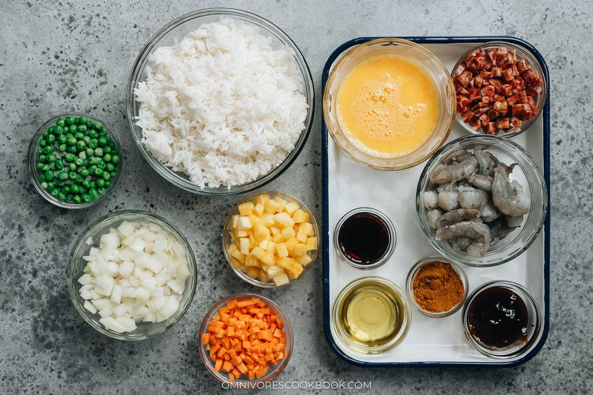 Ingredients laid out in bowls, including rice, shrimp, eggs, lap cheung, pineapple, peas, onion, carrot, and sauces.