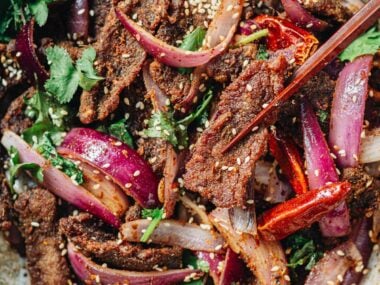 Chopsticks picking up a piece of cumin beef from a bowl filled with red onions and cilantro.