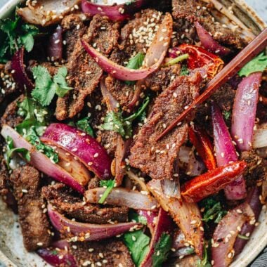 Chopsticks picking up a piece of cumin beef from a bowl filled with red onions and cilantro.