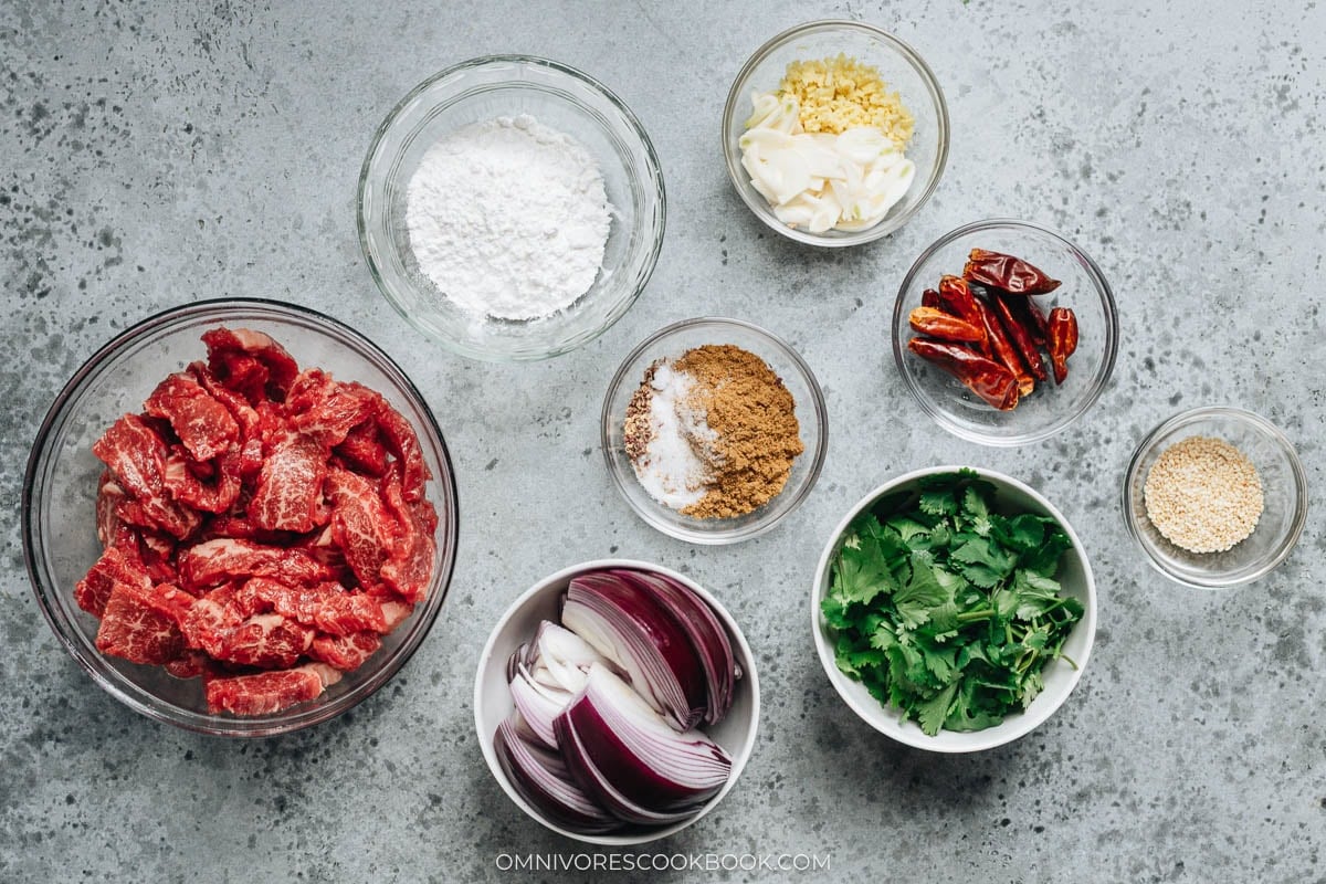 Overhead view of prepped ingredients including sliced beef, red onion, cornstarch, dried chilies, garlic, ginger, cumin spice mix, sesame seeds, and cilantro arranged in glass bowls.