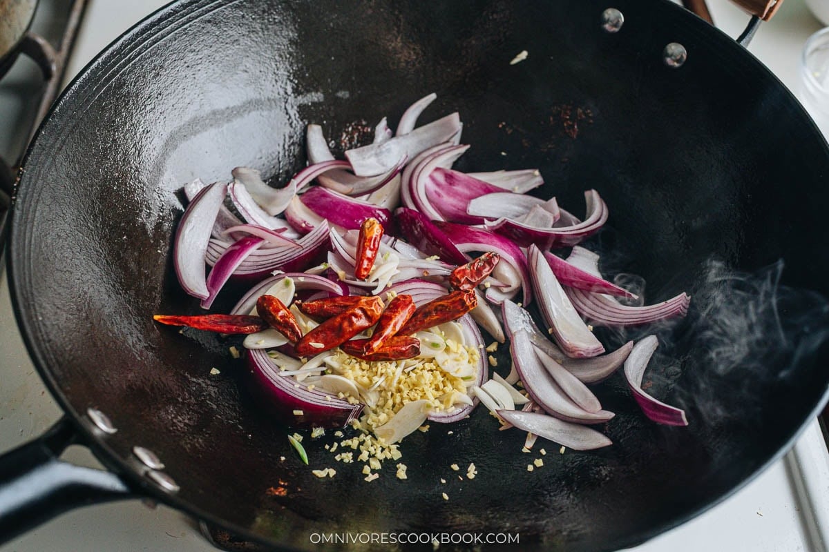 Aromatics including red onion, dried chilies, garlic, and ginger sizzling in a hot wok.