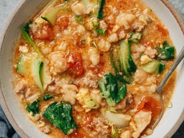 A bowl of Ge Da Tang served on a table, filled with chewy dough drops, wilted bok choy, ground pork, and soft egg ribbons in a tomato-based broth.