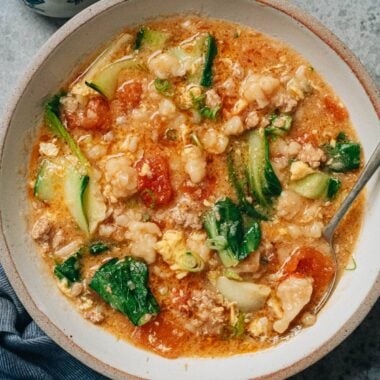 A bowl of Ge Da Tang served on a table, filled with chewy dough drops, wilted bok choy, ground pork, and soft egg ribbons in a tomato-based broth.