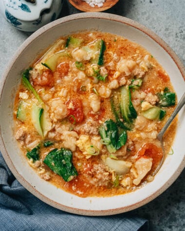 A bowl of Ge Da Tang served on a table, filled with chewy dough drops, wilted bok choy, ground pork, and soft egg ribbons in a tomato-based broth.