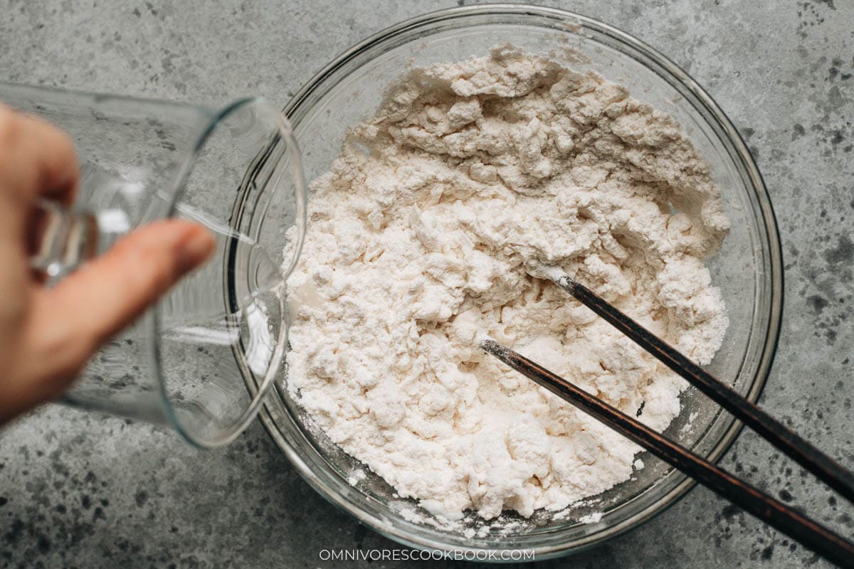 Pouring warm water into flour in a mixing bowl while stirring with chopsticks to create dough drops.