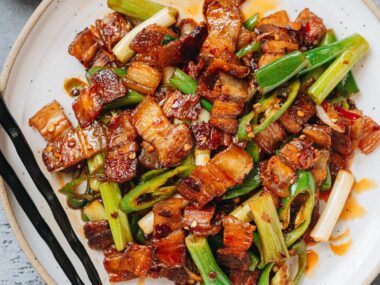 A plate of twice cooked pork with golden pork belly slices, green onions, and green chili peppers tossed in a glossy red sauce, served with black chopsticks.