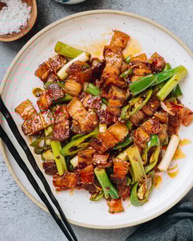 A plate of twice cooked pork with golden pork belly slices, green onions, and green chili peppers tossed in a glossy red sauce, served with black chopsticks.