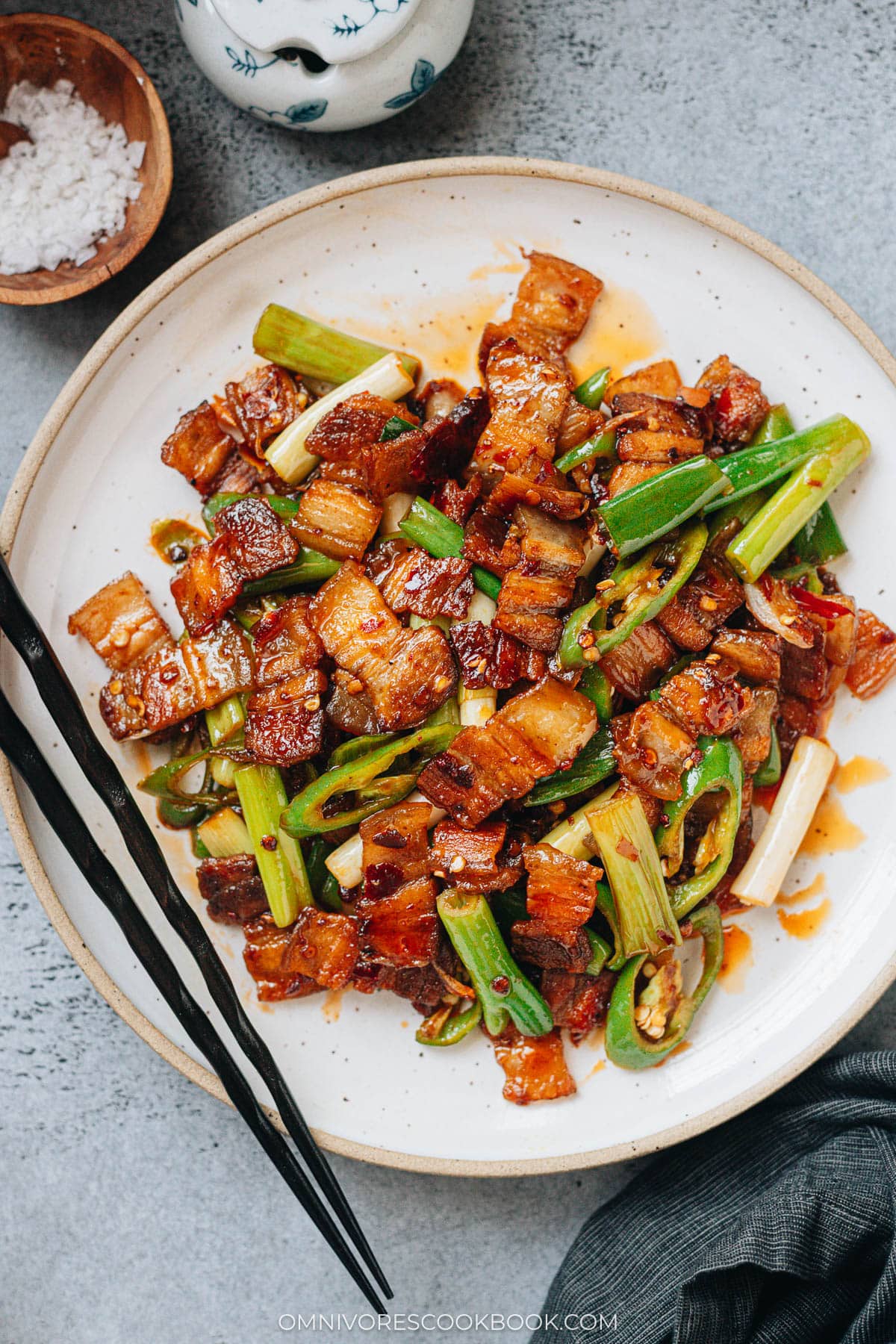 A plate of twice cooked pork with golden pork belly slices, green onions, and green chili peppers tossed in a glossy red sauce, served with black chopsticks.
