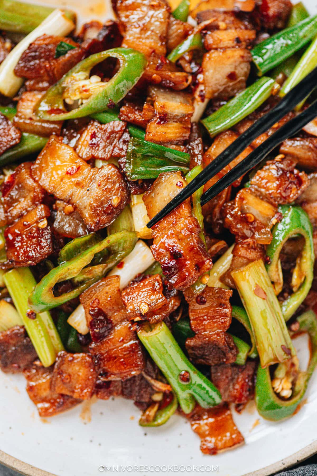 Close-up of pork belly slices in twice cooked pork, with chopsticks picking up a glossy piece surrounded by peppers and green onions.