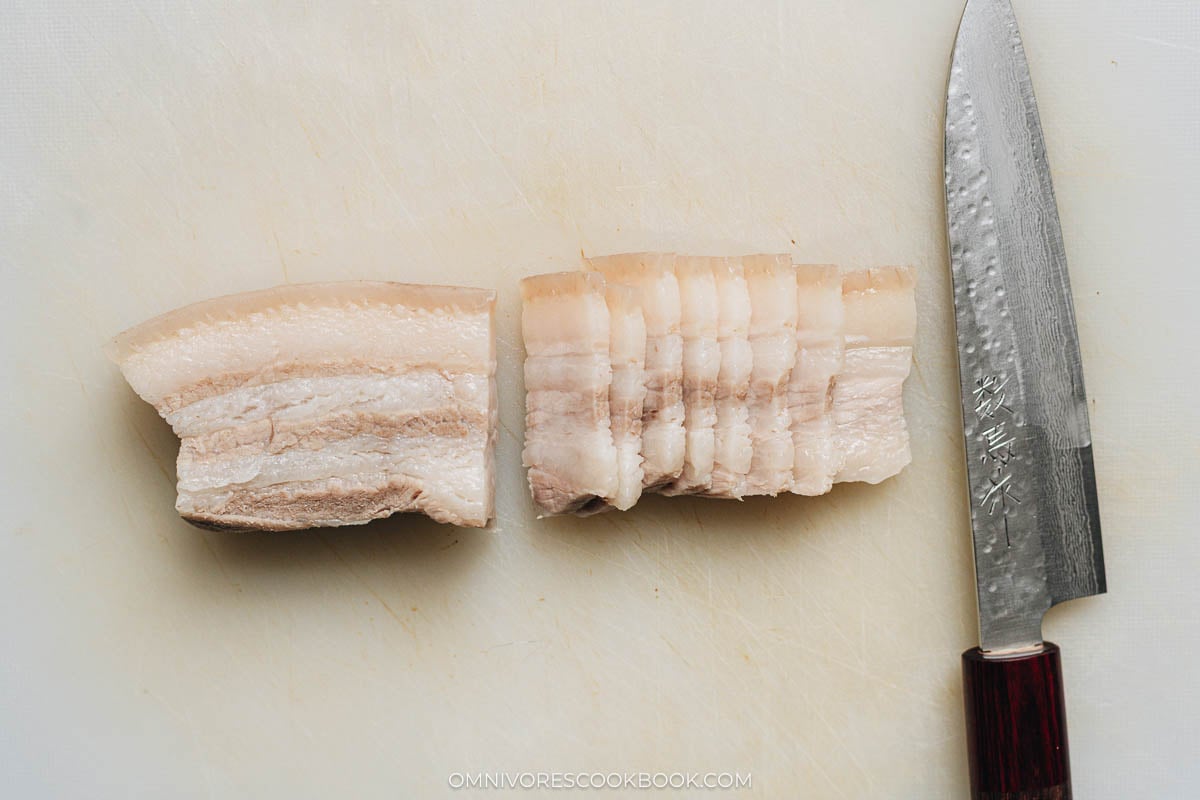 Par-cooked pork belly being sliced thinly on a cutting board.