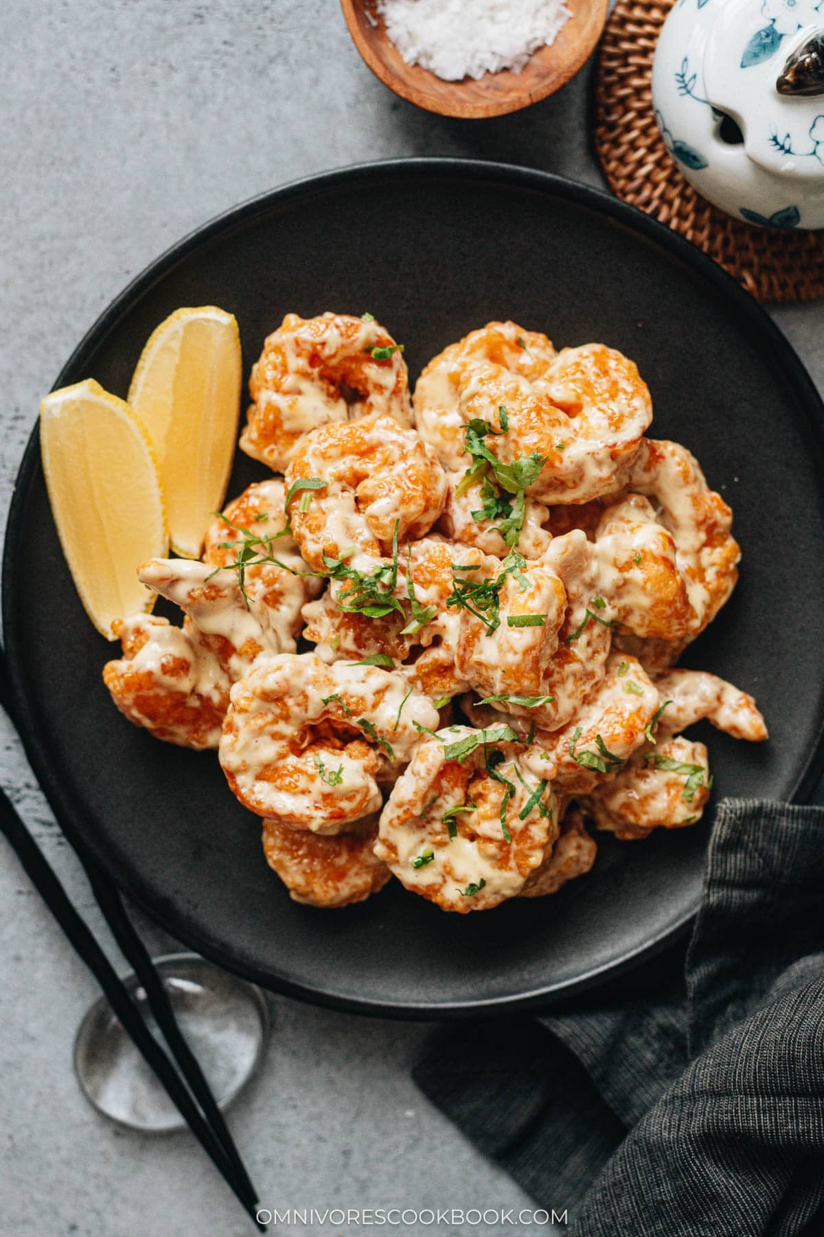 Wasabi mayo shrimp served on a black plate with chopped cilantro and lemon wedges.