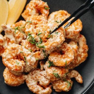 Closeup of a shrimp picked up with black chopsticks, showing the crisp coating and creamy wasabi mayo.