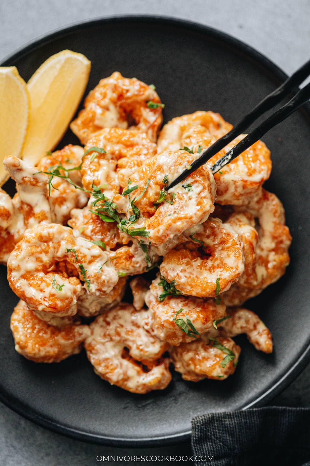 Closeup of a shrimp picked up with black chopsticks, showing the crisp coating and creamy wasabi mayo.