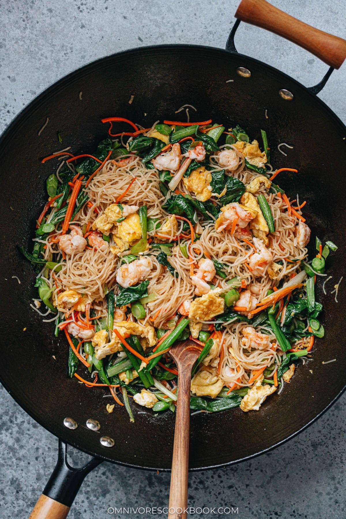 Shrimp mei fun stir fried in a wok with shrimp, egg, carrots, and choy sum.