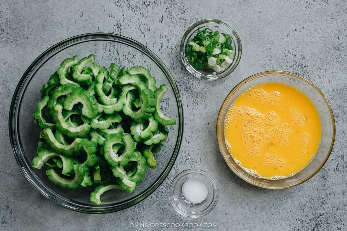 Sliced bitter melon with chopped green onions and beaten eggs on a gray surface.