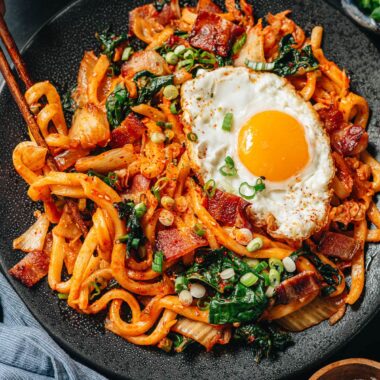 Bowl of bacon kimchi fried udon topped with a raw egg yolk, nori, and green onions