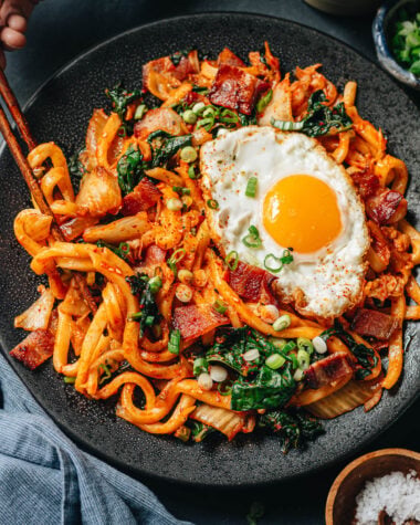 Bowl of bacon kimchi fried udon topped with a raw egg yolk, nori, and green onions