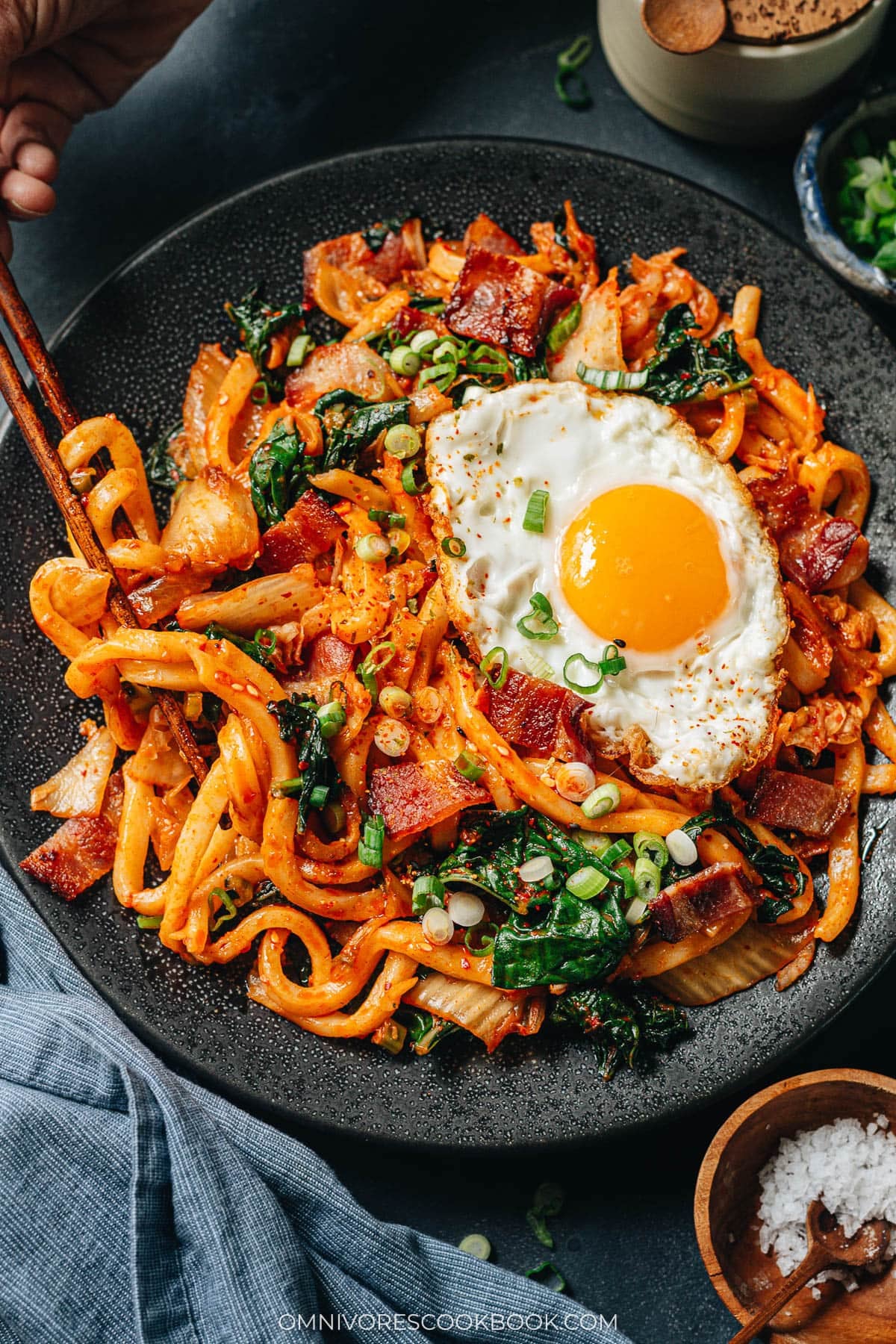 Bowl of bacon kimchi fried udon topped with a raw egg yolk, nori, and green onions