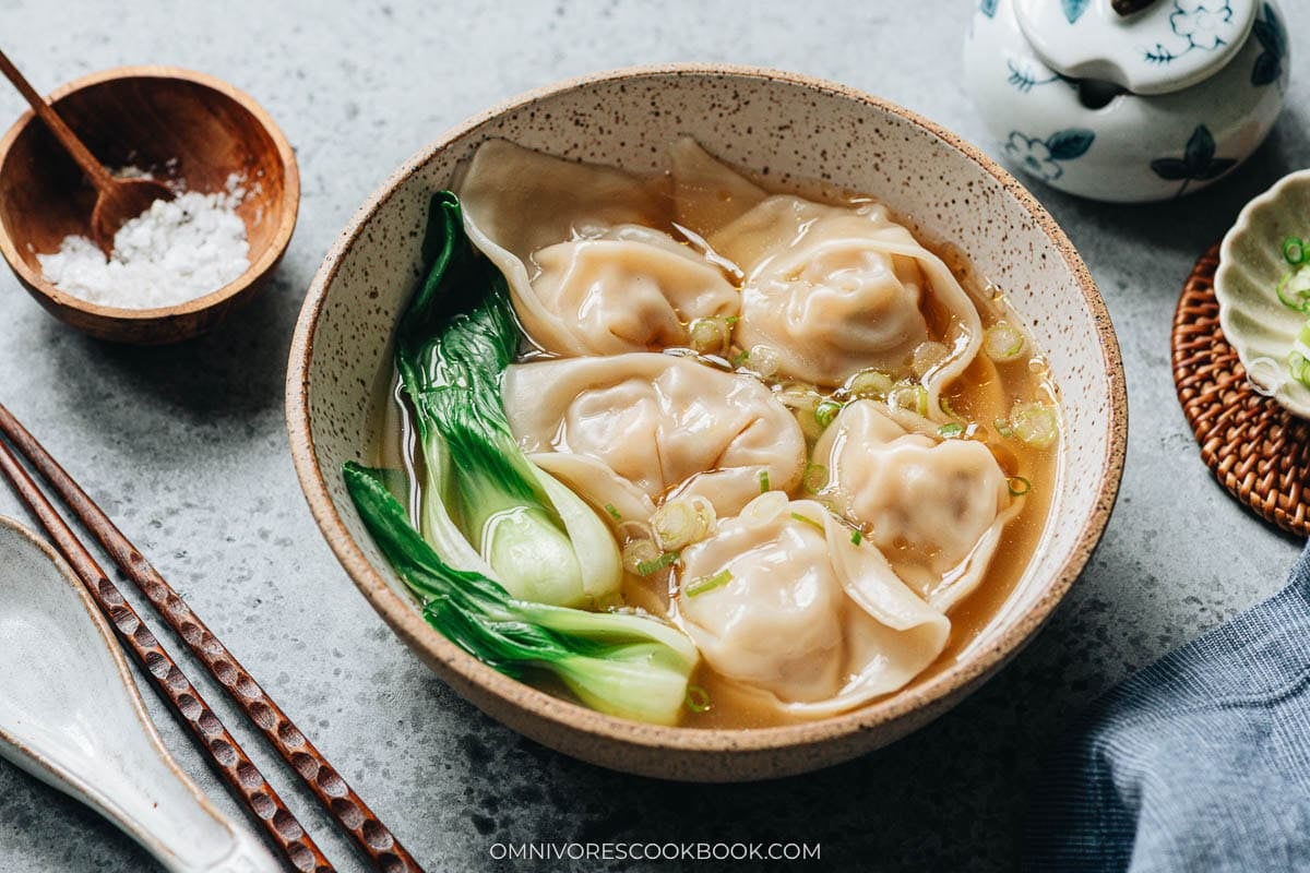 Assembling wonton soup, ladling hot broth over wontons and bok choy in a serving bowl
